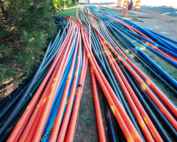 Dozens of orange, blue, and black utility conduits laid in parallel rows across a grass shoulder, converging toward a road construction site with orange and white safety barrels visible in the background.