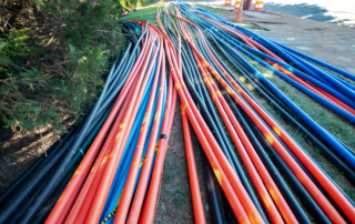 Dozens of orange, blue, and black utility conduits laid in parallel rows across a grass shoulder, converging toward a road construction site with orange and white safety barrels visible in the background.