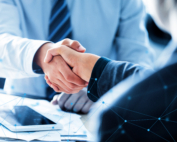 Two business professionals in suits shaking hands over a desk with documents and a tablet, overlaid with a blue digital network graphic.