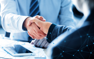 Two business professionals in suits shaking hands over a desk with documents and a tablet, overlaid with a blue digital network graphic.